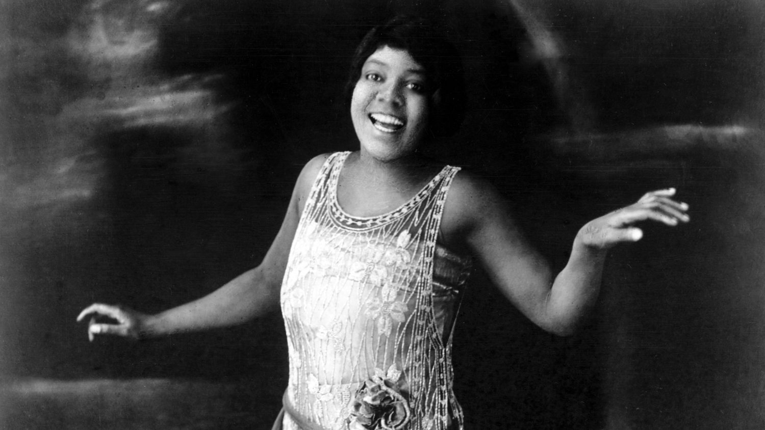 Bessie Smith, c. 1922, in a beaded sleeveless gown, arms extended in a stage pose, smiling toward the camera.