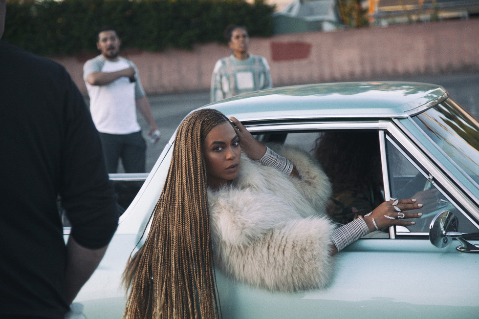 Beyoncé leaning out of the rear window of a vintage light-blue Chevrolet El Camino in an empty parking lot at dusk. She wears long honey-colored braids and a white fur coat over a striped grey sleeve. Two figures in the background watch from a distance.