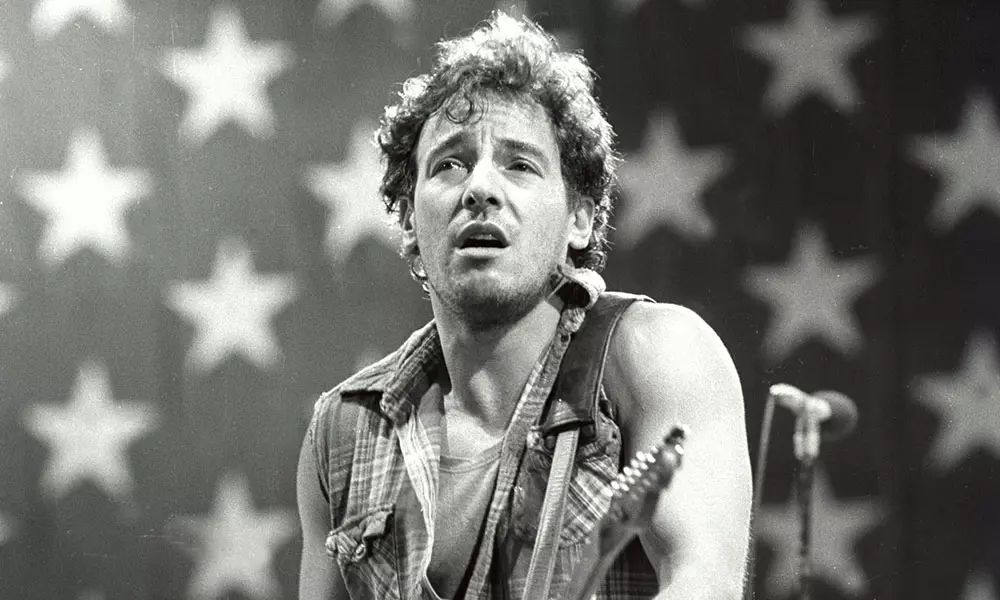 Black-and-white photograph of Bruce Springsteen mid-performance on stage in front of a large draped American flag backdrop with bold white stars on a darker field. He is shown from the waist up, wearing a sleeveless plaid button-up shirt with the sleeves cut away to expose his arms; a guitar strap crosses his chest. His curly hair falls down to his shoulders, and he is looking upward with his mouth slightly open, mid-vocal phrase. A microphone stand stands to his right at chest height.