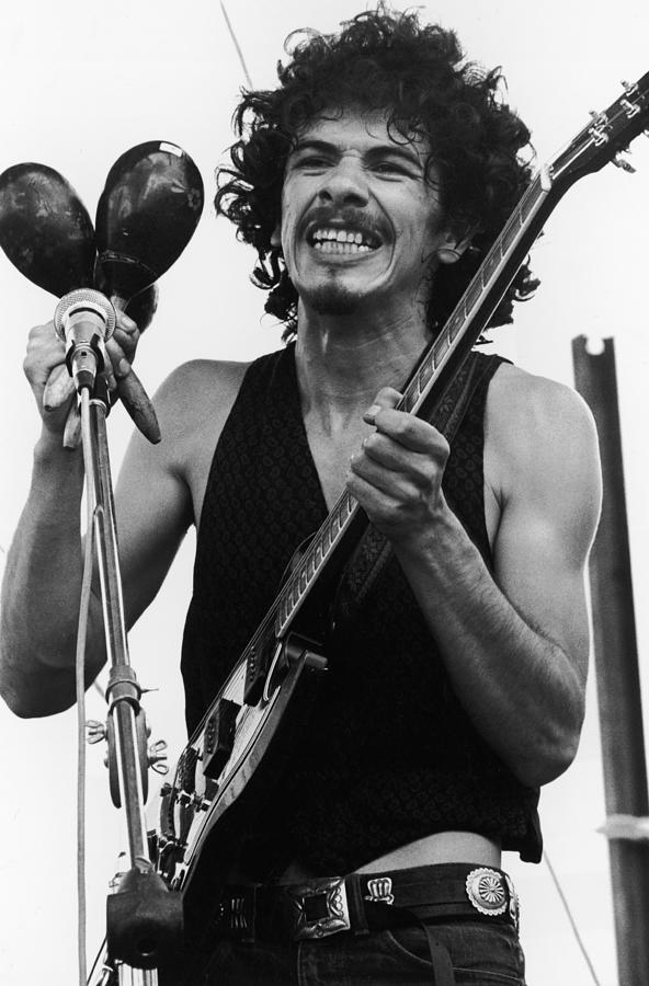 Black-and-white photograph of Carlos Santana onstage at the Woodstock Music Festival, August 16, 1969. Santana, in his early twenties, is shown from the waist up, wearing a sleeveless black knit vest, dark jeans, and a wide concho-style belt. He has a thick head of dark curly hair, a mustache and small goatee, and is grinning broadly. In his right hand he holds a pair of large dark maracas next to a vocal microphone on a boom stand; in his left hand he steadies the neck of a Gibson SG electric guitar.