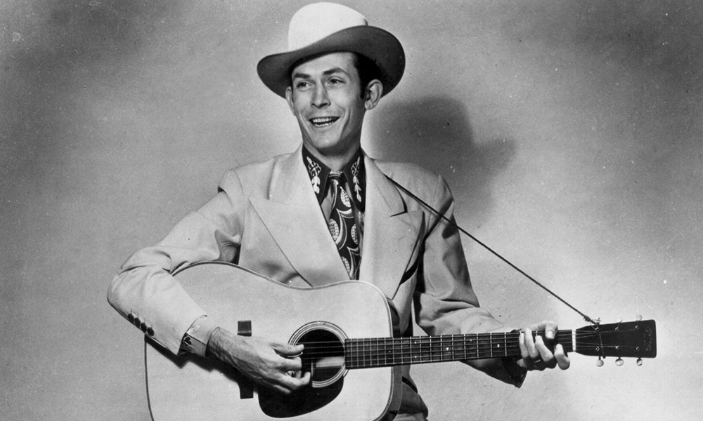 Hank Williams in a publicity portrait, holding an acoustic guitar, wearing a white Stetson hat and stage suit, smiling.