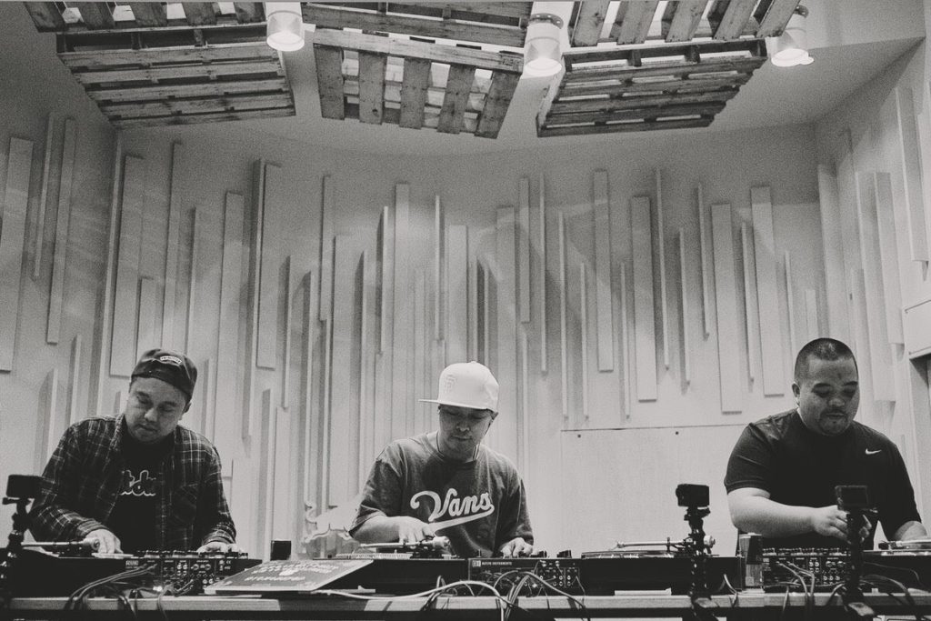 Black-and-white photograph of three DJs at three parallel turntable-and-mixer setups, viewed from the front. The studio is bright and modern, with white sound-diffusion paneling on the back wall and a wooden pallet treatment overhead. The DJ on the left wears a baseball cap and a plaid shirt; the middle DJ wears a white SF Giants cap and a black Vans baseball jersey; the DJ on the right wears a black Nike t-shirt. Each rig has a turntable on either side of a mixer, and small camera mounts are visible at the front of each station for filming the hand-work.