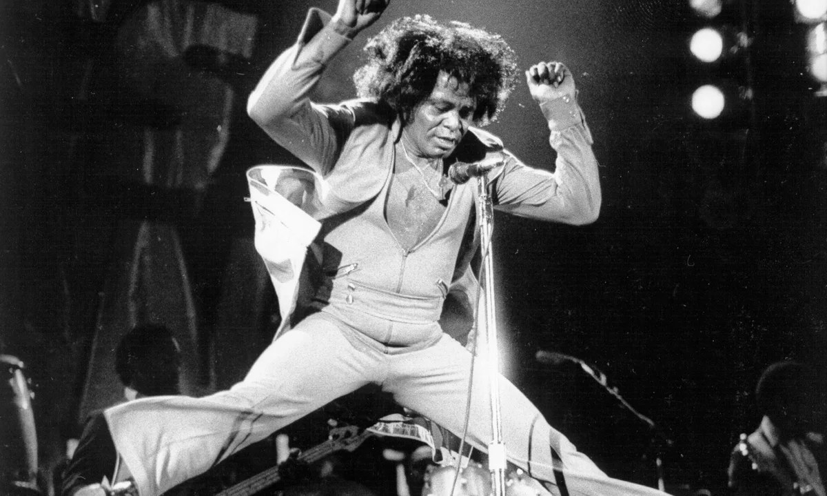 James Brown mid-air during a stage performance, both fists raised above his head, legs in a wide split, wearing a glittering jumpsuit with a cape over his shoulders. The band is partly visible behind him under stage lights.