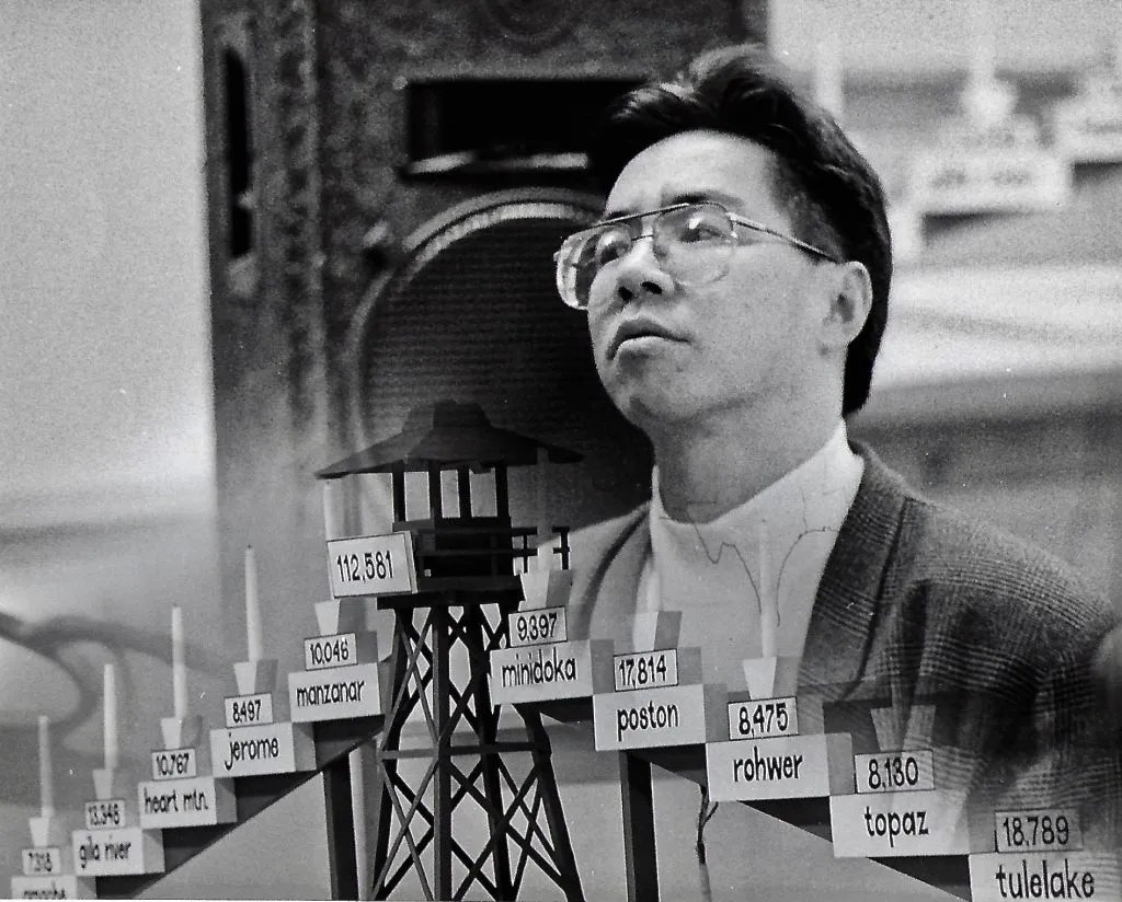 Black-and-white photograph of Jon Jang in three-quarter profile, head turned upward, wearing wire-rimmed eyeglasses, a dark jacket over a light shirt. Behind him, a model of a wooden guard tower bearing a plaque with the number 112,581 (the approximate total of Japanese Americans incarcerated during World War II). In the foreground, a row of small candle-name plaques runs across the bottom of the image, each bearing the name and approximate incarceree count of one of the ten WRA concentration camps: amache 7,318; gila river 13,348; heart mtn. 10,767; jerome 8,497; manzanar 10,046; minidoka 9,397; poston 17,814; rohwer 8,475; topaz 8,180; tulelake 18,789.