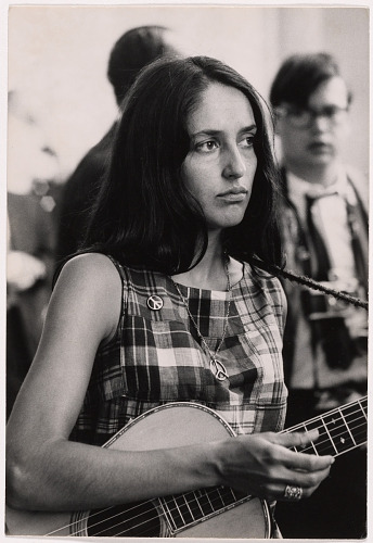 Black-and-white photograph of Joan Baez at the August 1963 March on Washington for Jobs and Freedom. She is photographed from the chest up, looking past the camera with a serious, composed expression. Her long dark hair falls past her shoulders. She wears a plaid sleeveless top with a peace-symbol pin attached at the chest and a thin chain necklace with a peace-symbol pendant. The neck of a guitar she is holding is visible in front of her at the bottom of the frame, with strings and tuning pegs in view. Two younger people are out of focus behind her: a man in a white shirt at her left shoulder, and a young man in glasses and a white shirt with a dark tie at her right. The background is a softly lit interior.