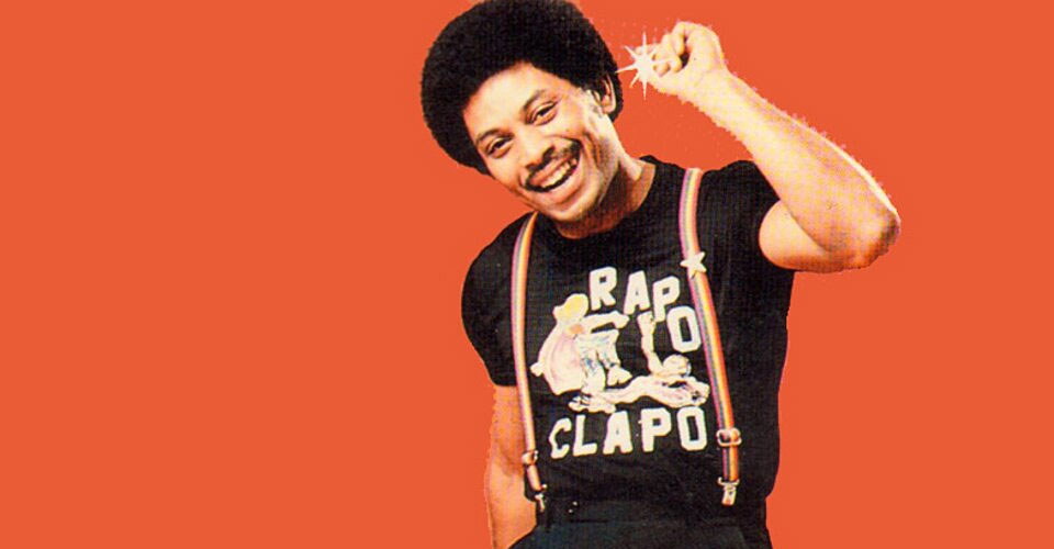Color photograph of Joe Bataan in his late thirties against a bright orange background, smiling broadly, with a large afro and mustache, snapping the fingers of his raised right hand. He wears a black t-shirt with a cartoon graphic that reads 'RAP-O CLAP-O,' and bright orange suspenders.
