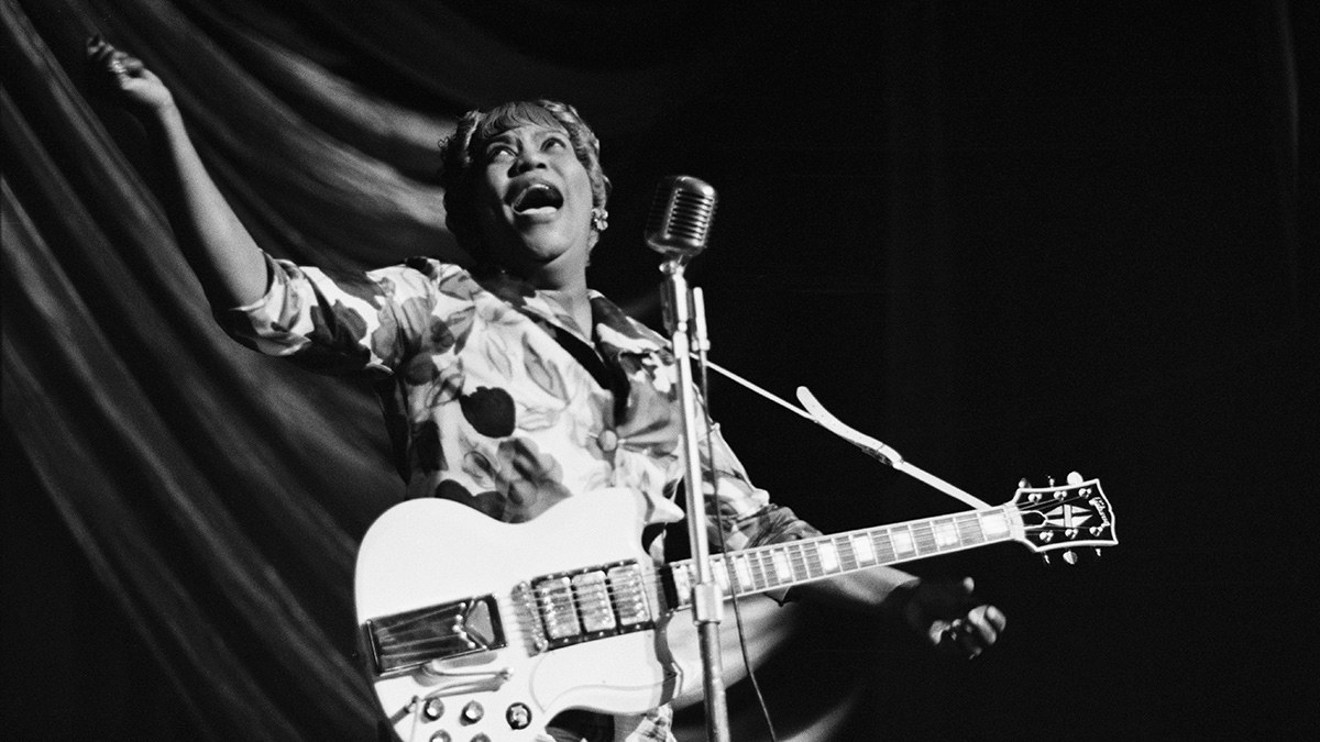 Sister Rosetta Tharpe in mid-performance, head tilted back and mouth open mid-phrase, left arm extended to her side in an open gesture, a white Gibson SG Custom electric guitar at her hip, in front of a vintage ribbon microphone and a dark stage curtain.