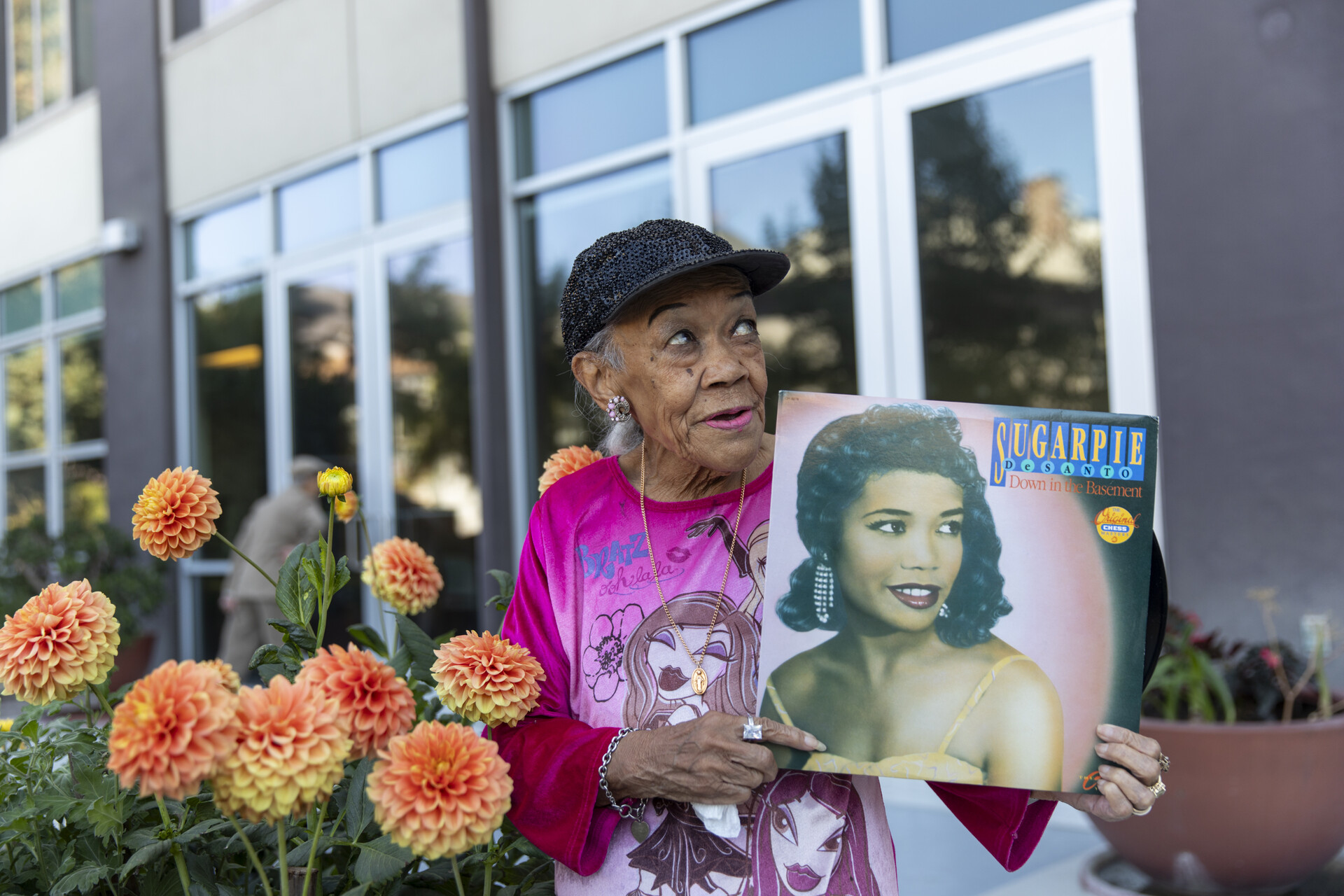 Sugar Pie DeSanto in 2023, holding her record Down in the Basement outside her Oakland home, with orange dahlias in the foreground.