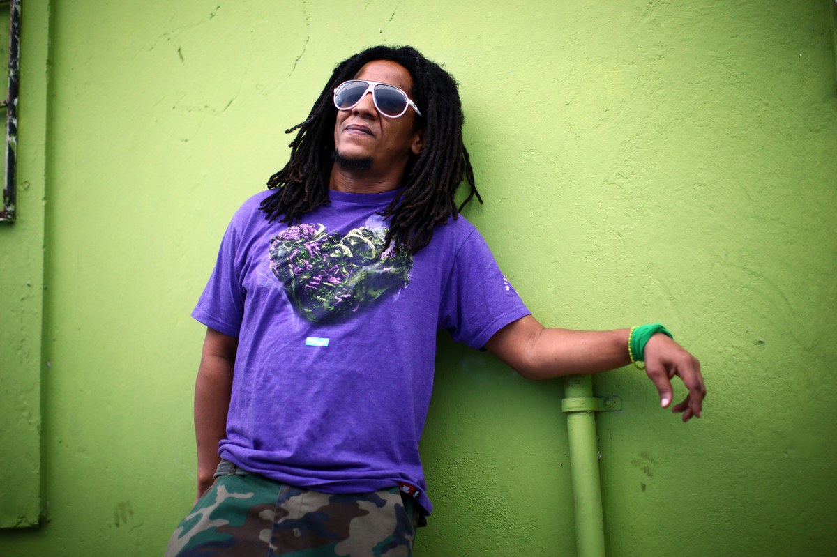 Documentary portrait of Tego Calderón outside his recording studio, El Sitio, in the Santurce neighborhood of San Juan, Puerto Rico. Tego, in his early forties, is shown from the chest up against a textured grey-brown urban wall. He wears a black hoodie with a white graphic on the chest, layered gold chains, and large dark aviator-style sunglasses. His long dreadlocks fall past his shoulders. The framing is candid and natural, the lighting is daylight, the mood is grounded rather than glamorous.