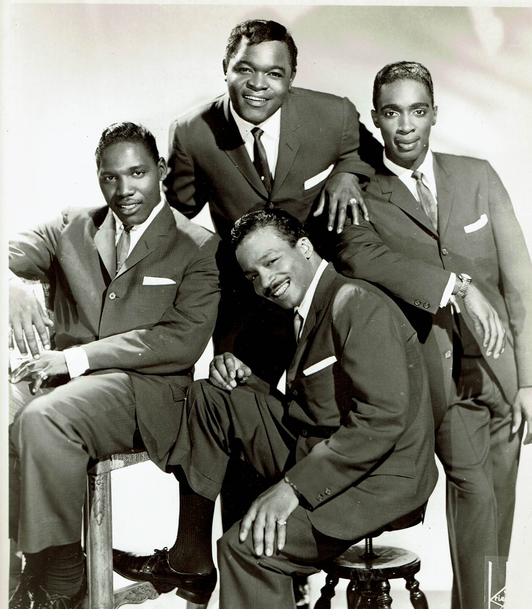 Black-and-white promotional studio photograph of four Black men in dark suits with white pocket squares and ties, arranged in a casual stacked group portrait. One man sits in the front center on a low piano stool, leaning forward with his hands folded over one knee and a wide moustachioed smile; the other three are clustered behind him. The man at the back stands tallest and grins broadly, looking down at the camera. A second man sits on the left with one elbow on a chair-arm. A third stands on the right with one hand resting on the seated man's shoulder, wearing a wristwatch and signet ring. The photographer's signature 'Kriegsmann' appears in white script at the lower-right corner of the print.