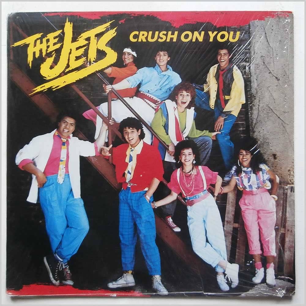 Color photograph of the original 1986 MCA Records 7-inch sleeve for The Jets, "Crush On You." Stylized yellow brushstroke logo reading THE JETS in the upper-left corner over a dark background; the title CRUSH ON YOU runs across the top in white. The eight Wolfgramm siblings, ages roughly 12 to 20, are arranged on a graffiti-marked metal staircase in mid-1980s pop fashion: pastel button-up shirts, bright bandanas, skinny ties in primary colors, rolled-up plaid pants, high-top sneakers and Converse low-tops, oversized blazers in red and white. The four older siblings are seated on the stairs and lean over a metal railing in the upper half of the image; the four younger siblings stand in front of the staircase in the lower half, smiling and posed mid-step.