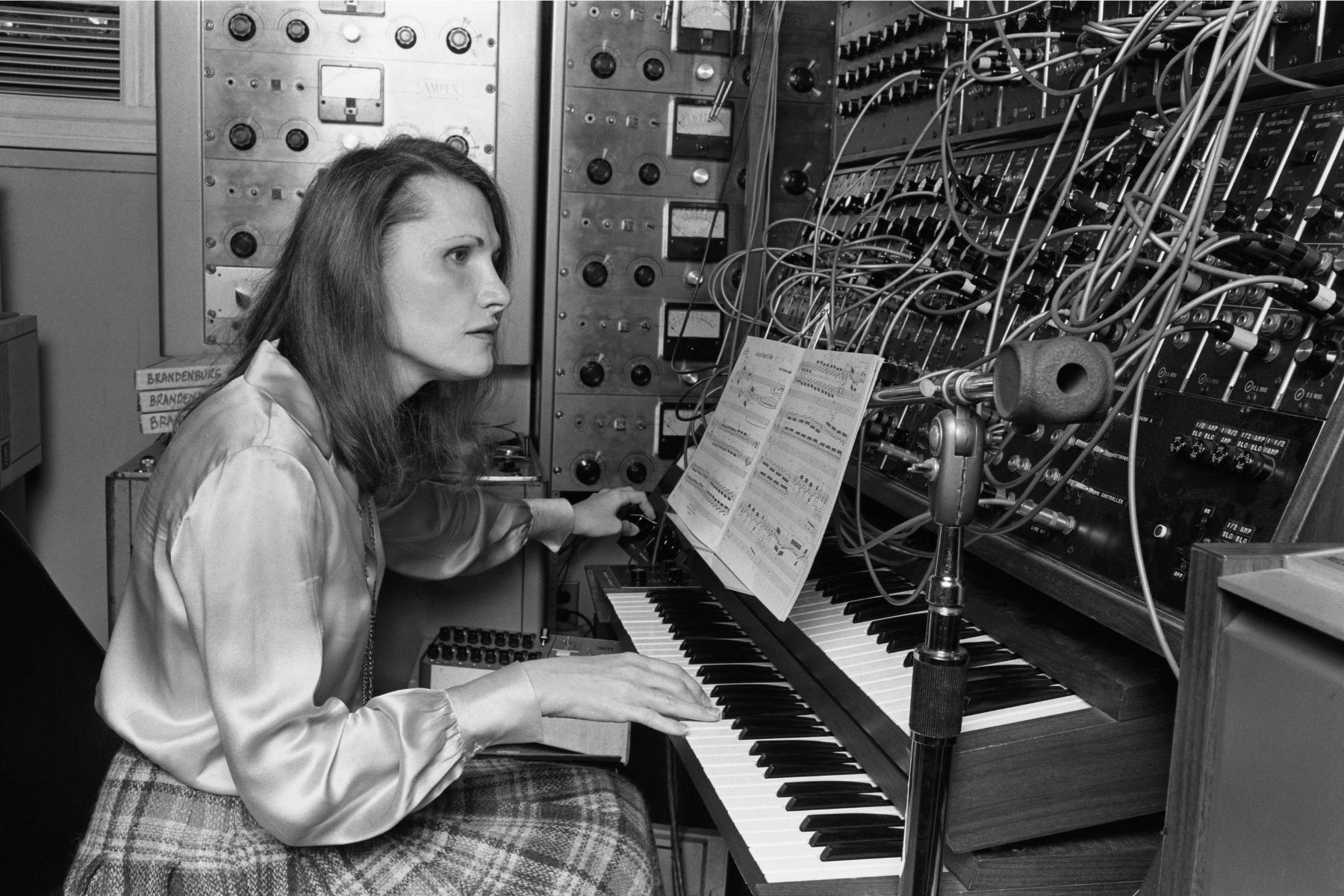 Black-and-white studio photograph of Wendy Carlos in profile, seated at a keyboard with both hands on the keys, looking up at a multi-page music score on a stand above the keyboard. She has shoulder-length dark hair and is wearing a satin blouse and a plaid skirt. To her left, against the wall, a rack-mounted Ampex tape machine and additional outboard audio equipment fill the studio wall from floor to ceiling, and a shelf at her left elbow carries three white tape boxes labeled BRANDENBURG, BRANDEN, BRA. To her right, a large Moog modular synthesizer fills the right side of the frame with rows of knobs, jacks, and patch cords running across the front panels; R.A. Moog module labels are visible on the right edge. A microphone on a boom stand stands between Carlos and the synthesizer, pointing at a small studio monitor speaker to the right of the keyboard.