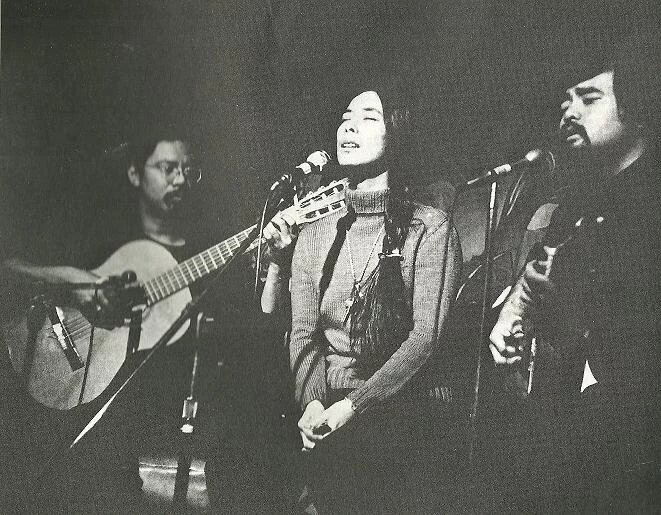 Black-and-white photograph of the three musicians performing together. Chris Iijima at left plays an acoustic guitar, head down toward his fingers, glasses on, dark shirt. Nobuko Miyamoto stands at the center microphone with her eyes closed mid-phrase, wearing a cream sweater and a long pendant. Charlie Chin at right plays a second acoustic guitar, mustache, dark shirt. Three vocal microphones and one instrument microphone are visible.