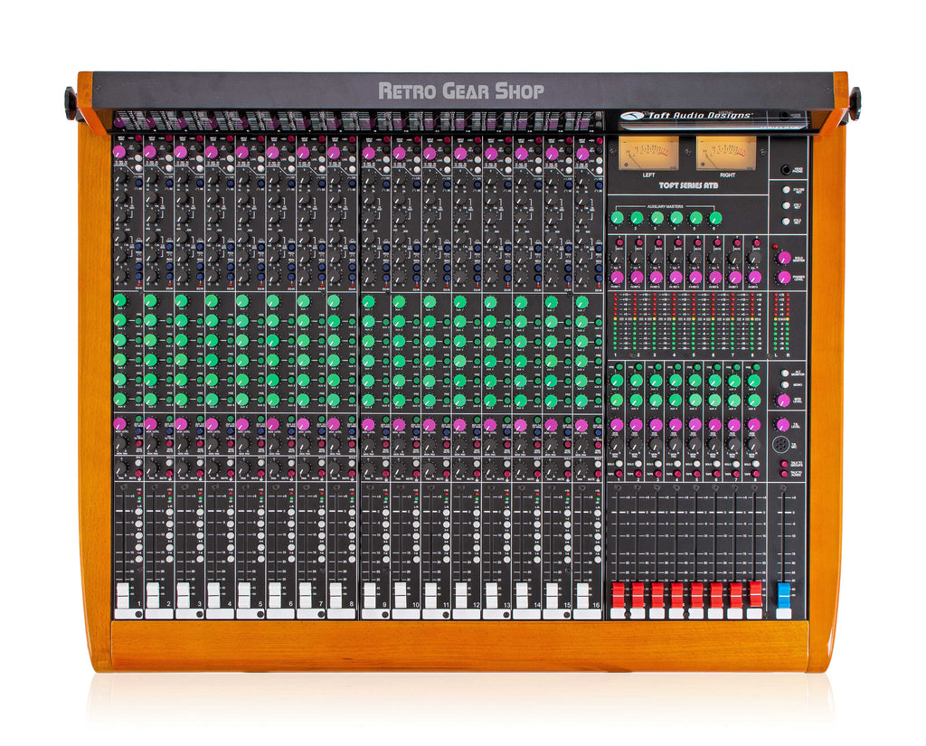 The Toft Audio Designs Series ATB 16-channel console, photographed from directly above. On the left two-thirds: sixteen identical input channel strips, each with rows of magenta, green, and pink knobs and a white vertical fader at the bottom. In the middle-right: eight narrower submaster strips with green knobs and red vertical faders. On the right: the master section with bargraph meters, the auxiliary master knobs, two VU meters, talkback controls, and the blue stereo master fader. The console sits in a warm orange wood frame.